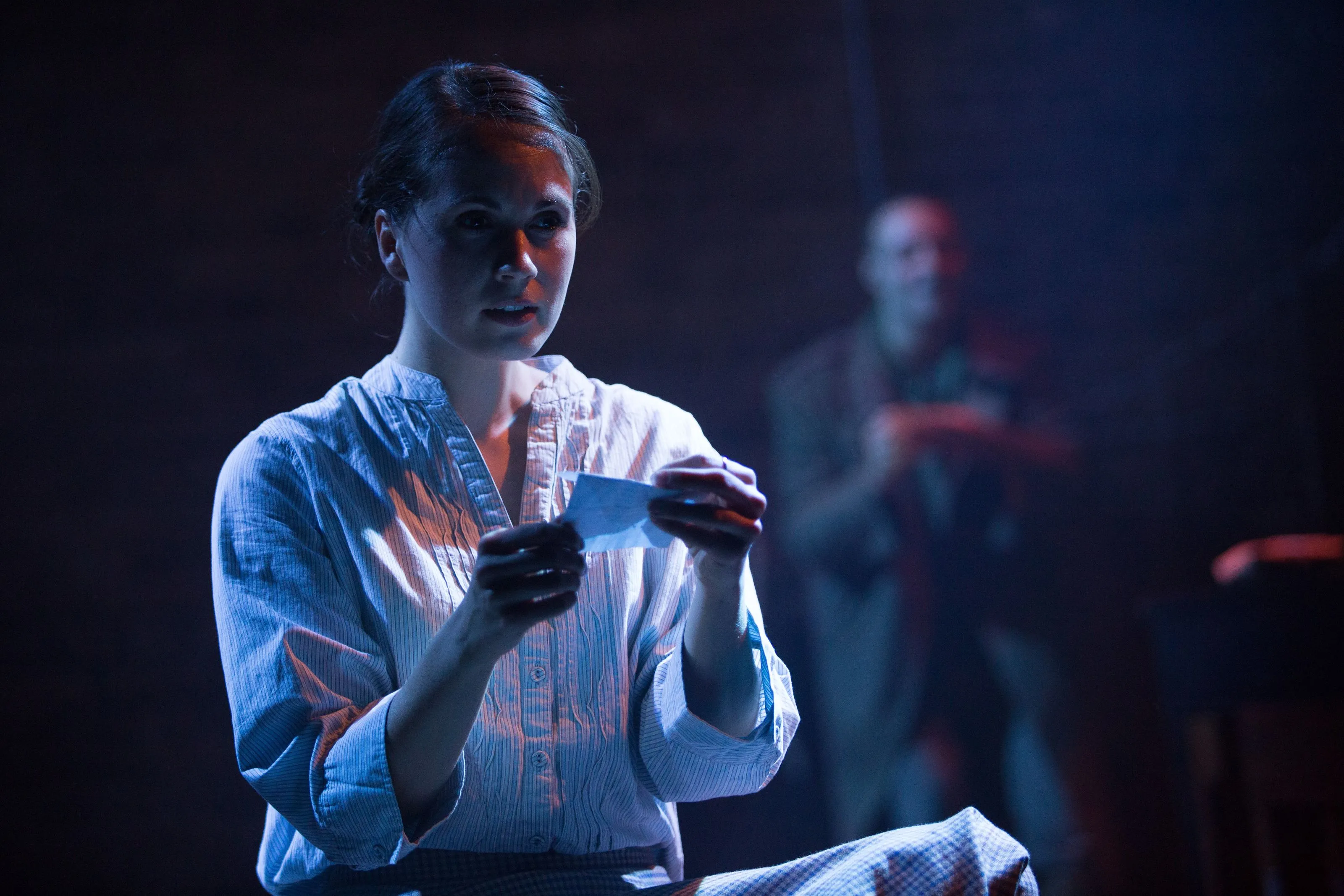 A steel blue light illuminates a woman as she reads from a slip of paper