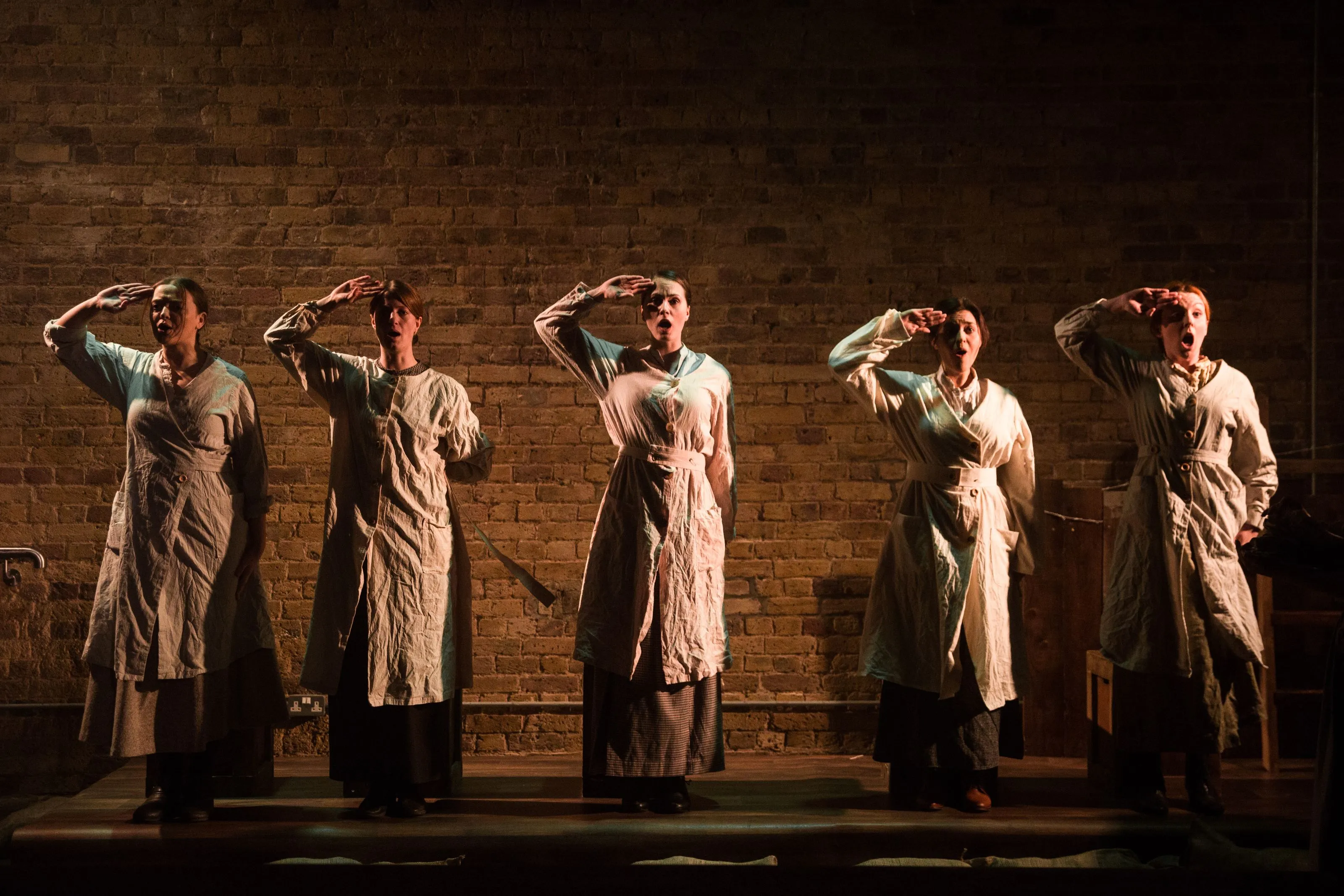 Five brightly side lit women salute in front of a brick wall