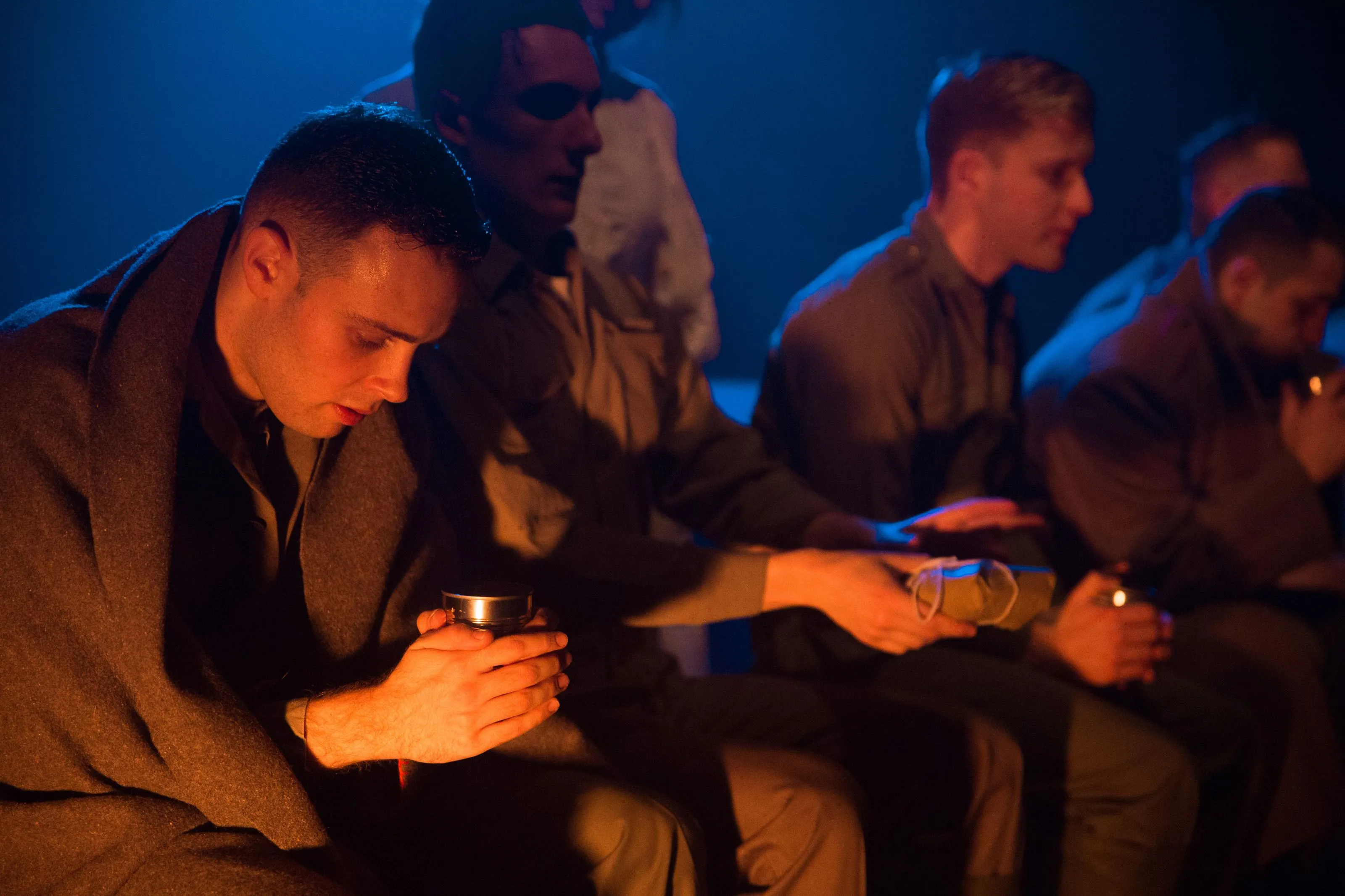 Actors dressed as soldiers huddle in a deep orange light with a cold dark backdrop