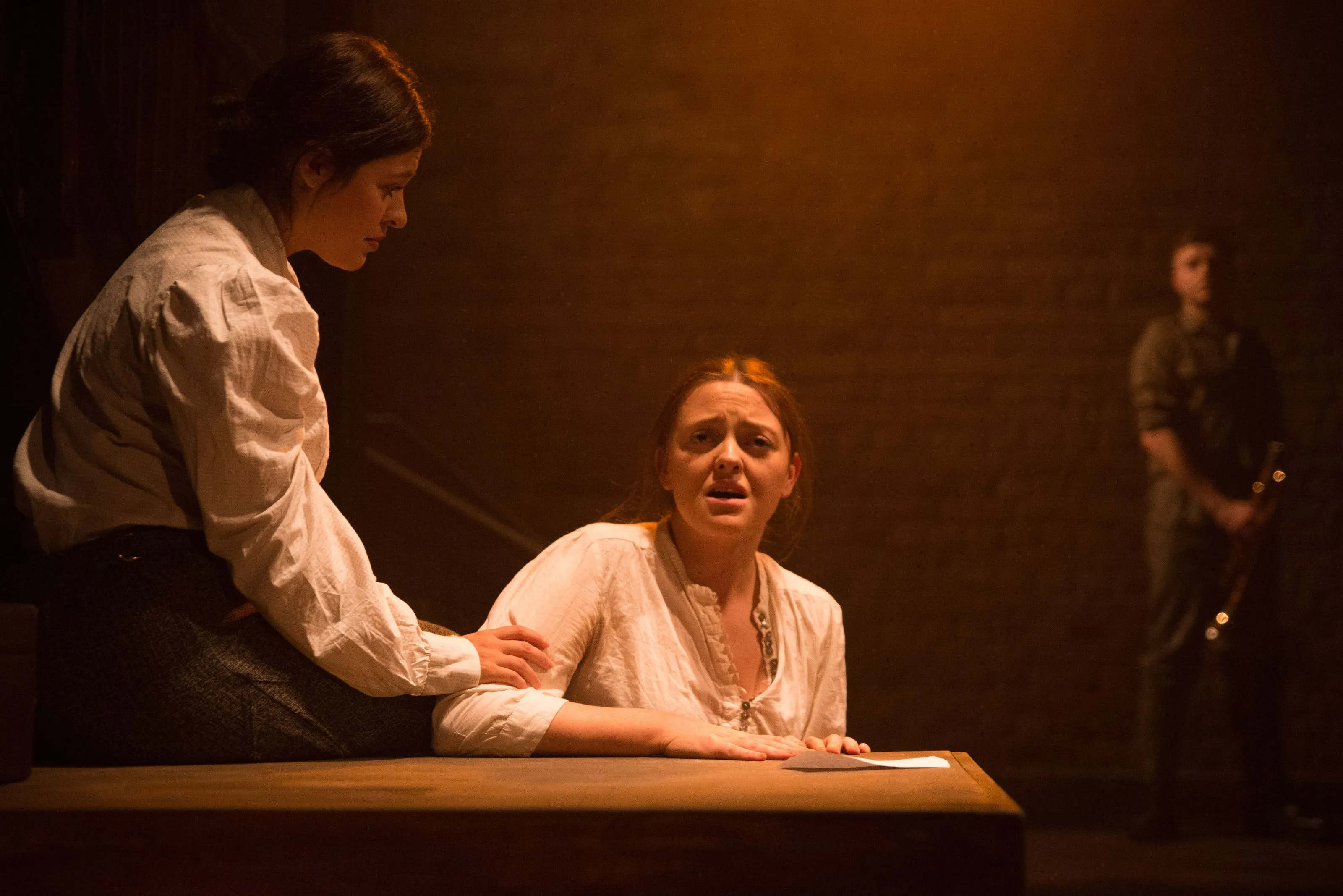 One woman consoles another at a table in a warm interior light