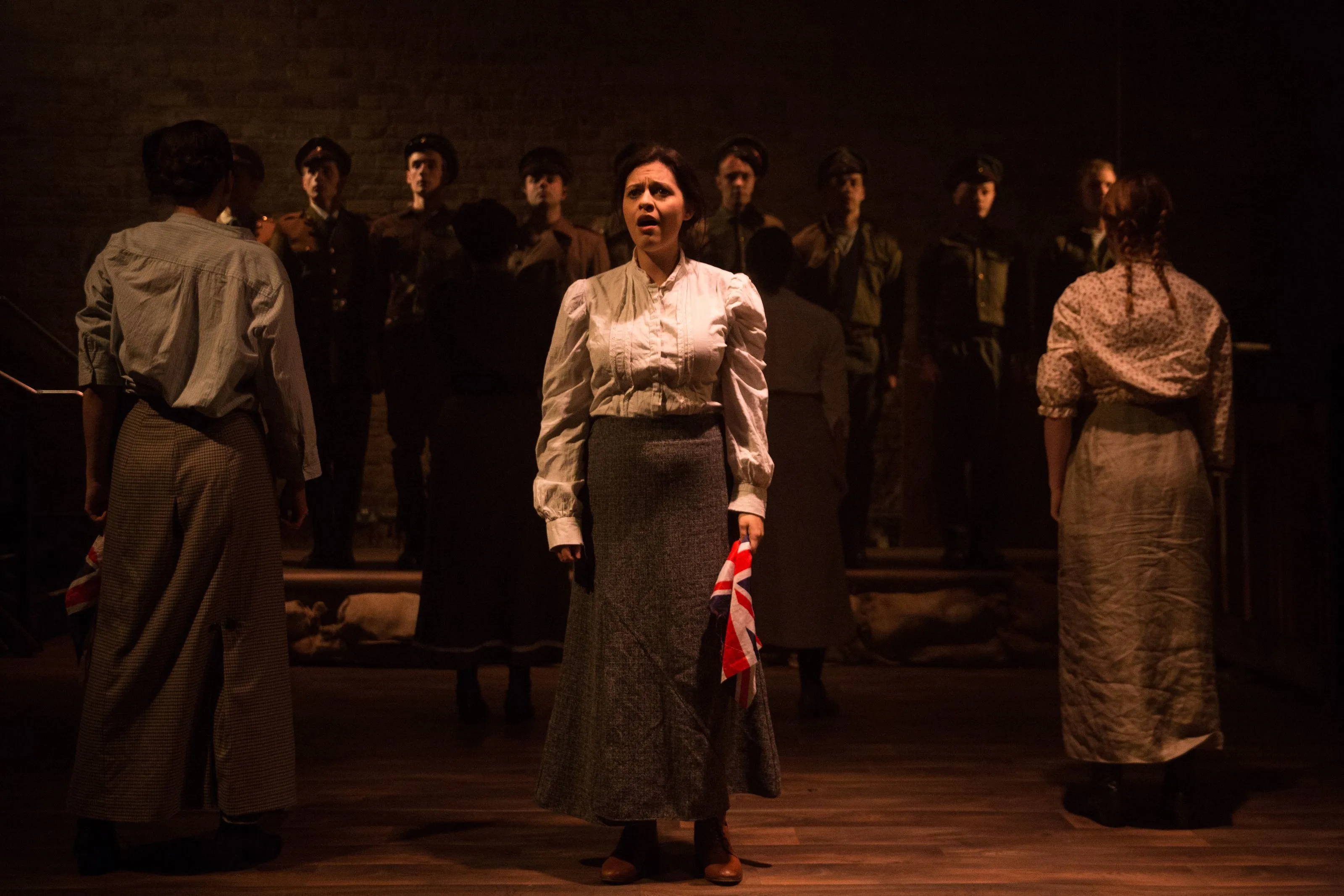 A distressed woman holding a Union Jack is spotlit onstage surrounded by other actors in shadow