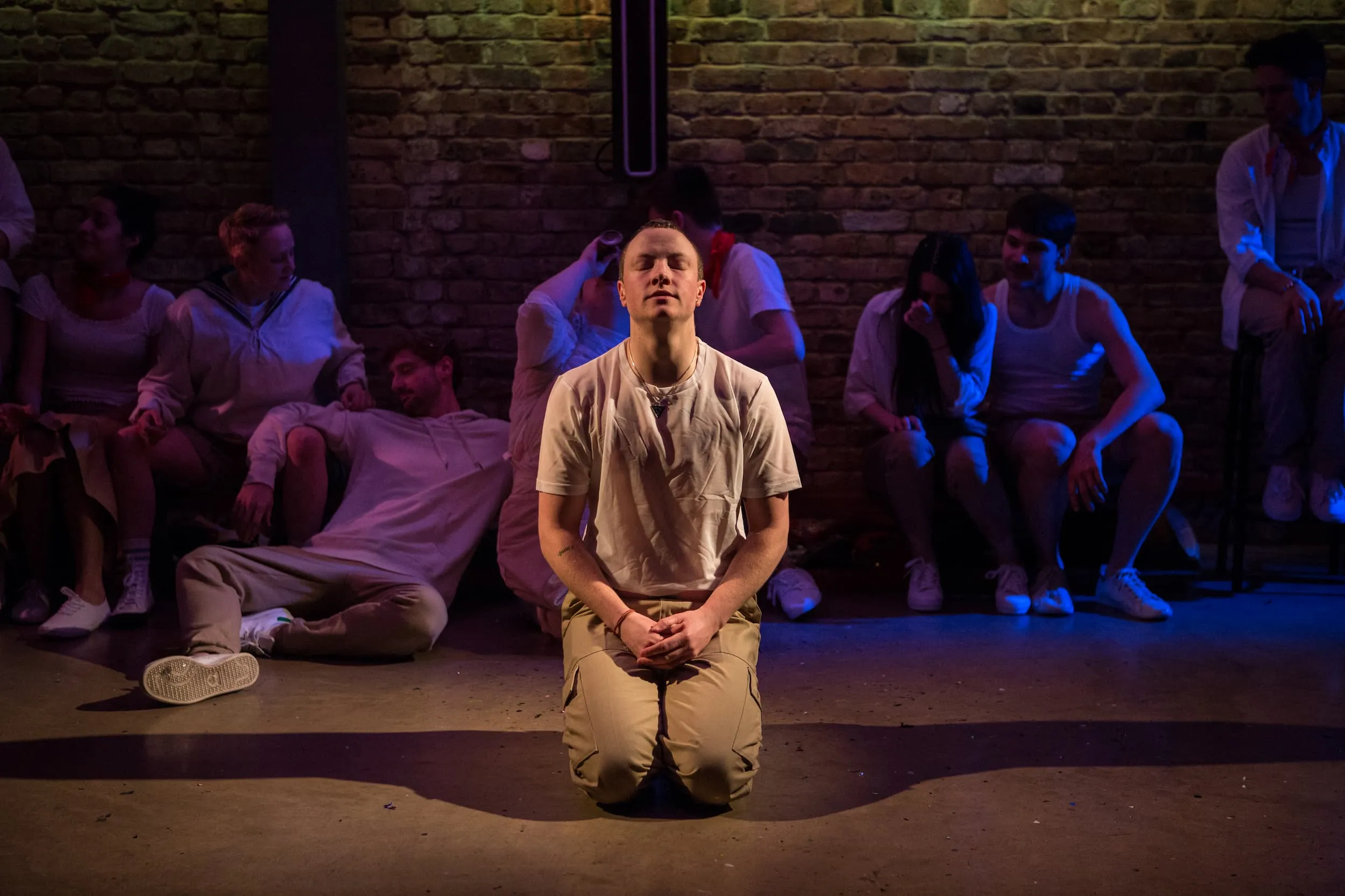 A man in a white t-shirt and khakis kneels with hands clasped and eyes closed in reflection.