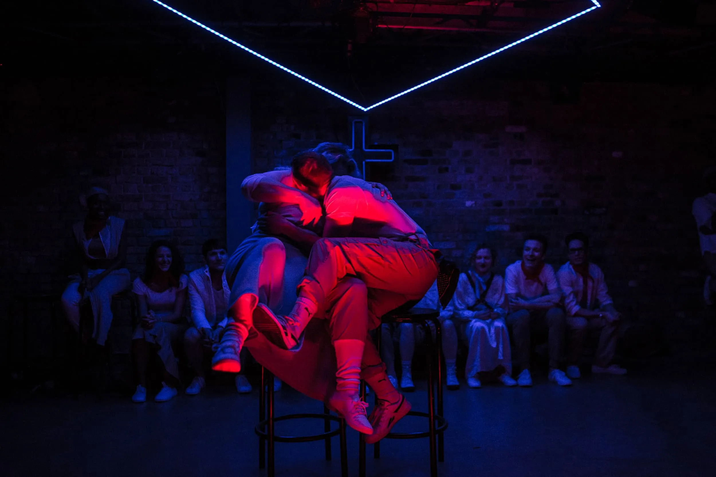 Two people embrace on bar stools in intense magenta light against a dim blue wash.