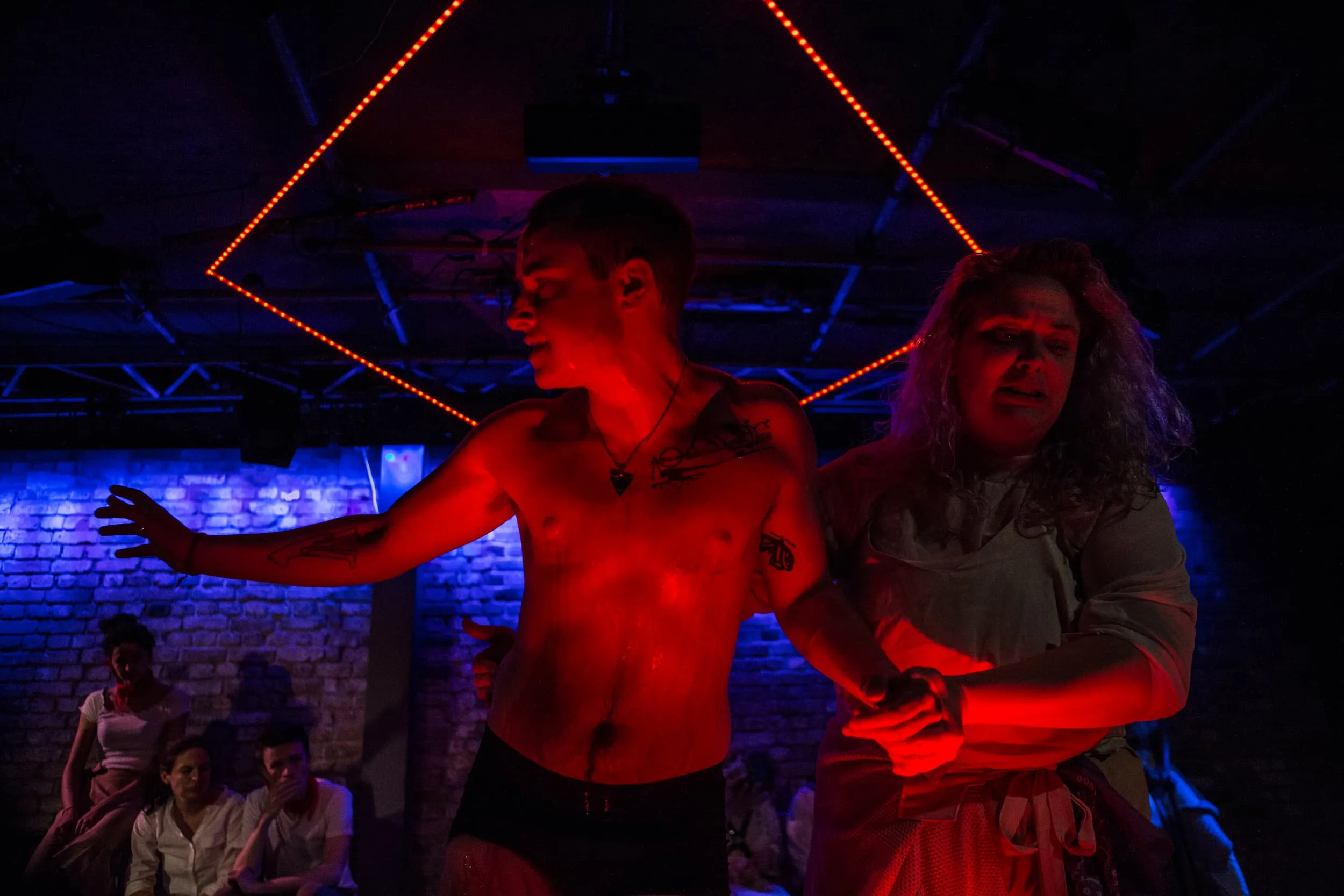 Under intense red light, a nearly naked man, dripping wet, is led away by a woman with long hair.