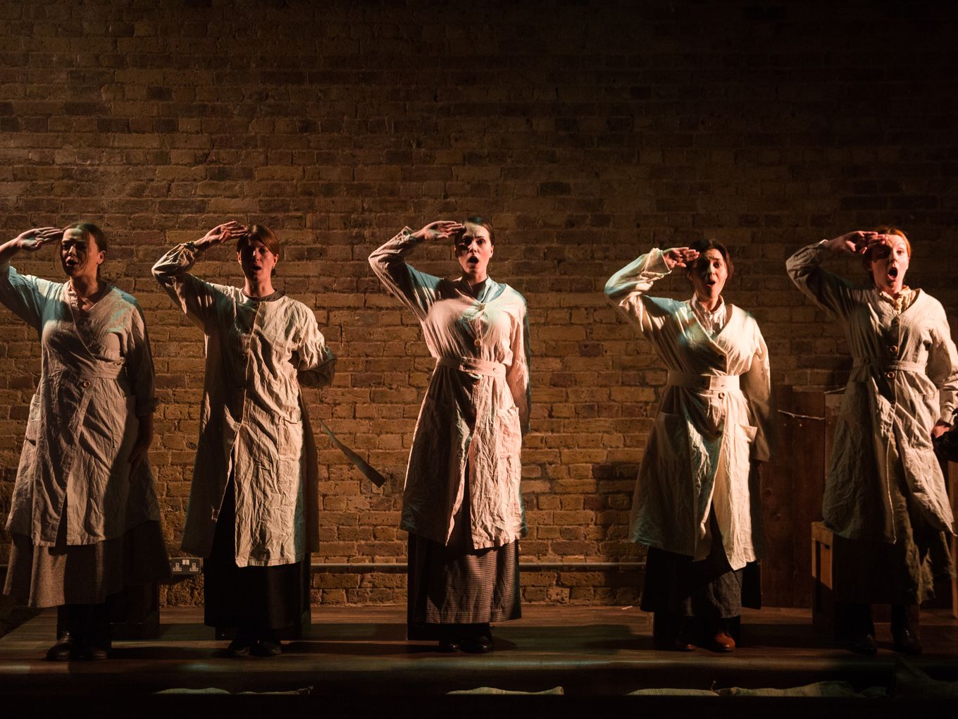 Five brightly side lit women salute in front of a brick wall