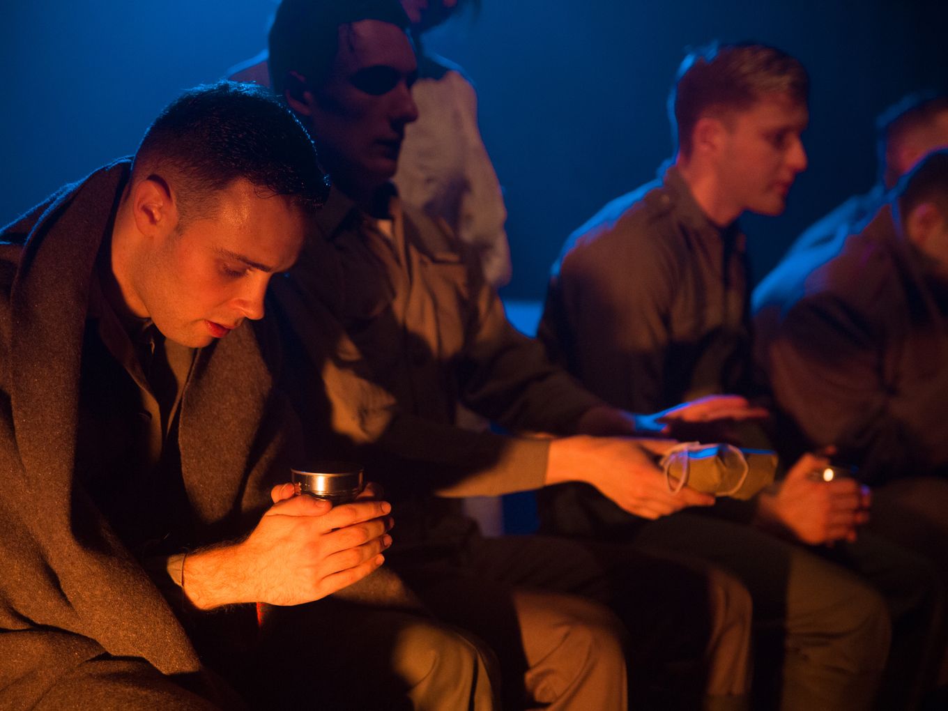 Actors dressed as soldiers huddle in a deep orange light with a cold dark backdrop