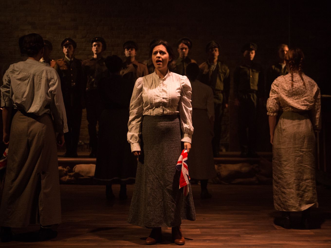 A distressed woman holding a Union Jack is spotlit onstage surrounded by other actors in shadow
