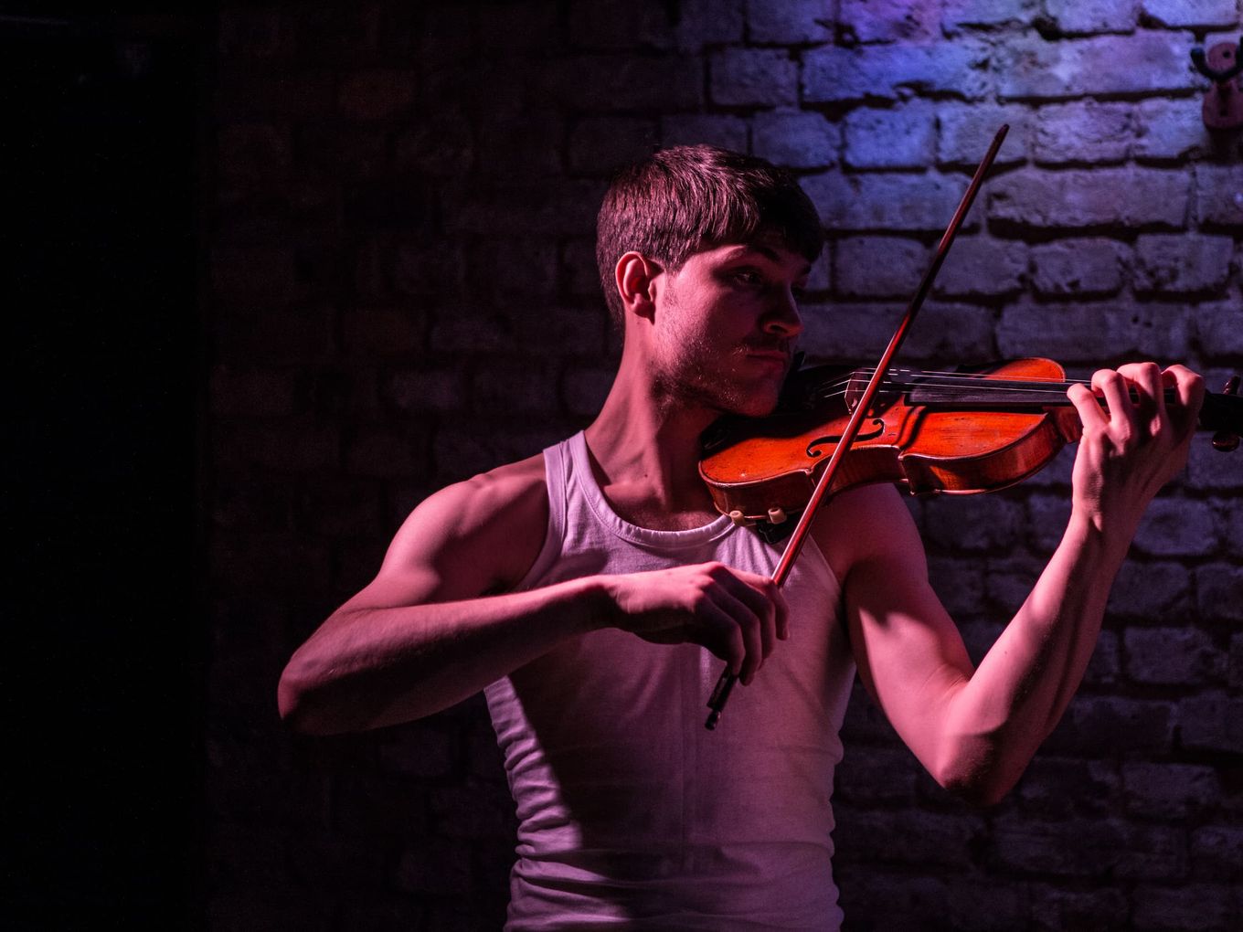 A man in a vest plays the violin in dim warm light