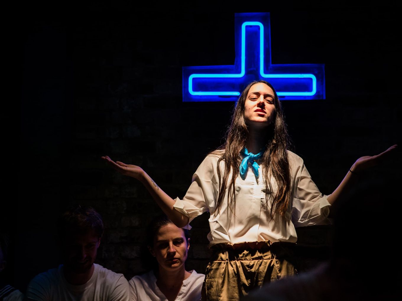 A person with long hair stands hands outstretched in front of a blue neon cross