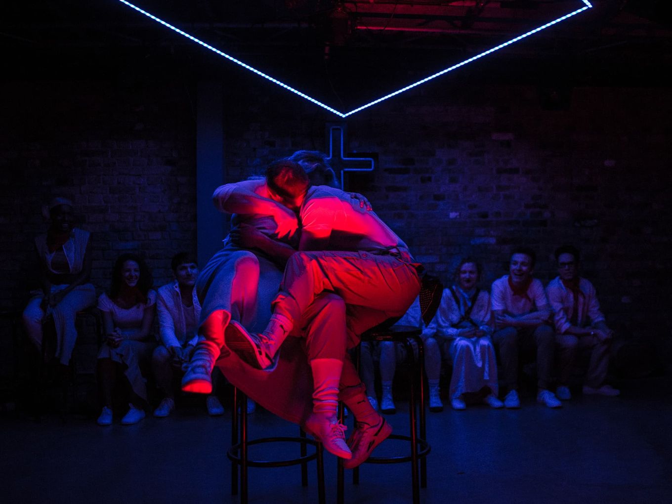 Two people embrace on bar stools in intense magenta light against a dim blue wash.
