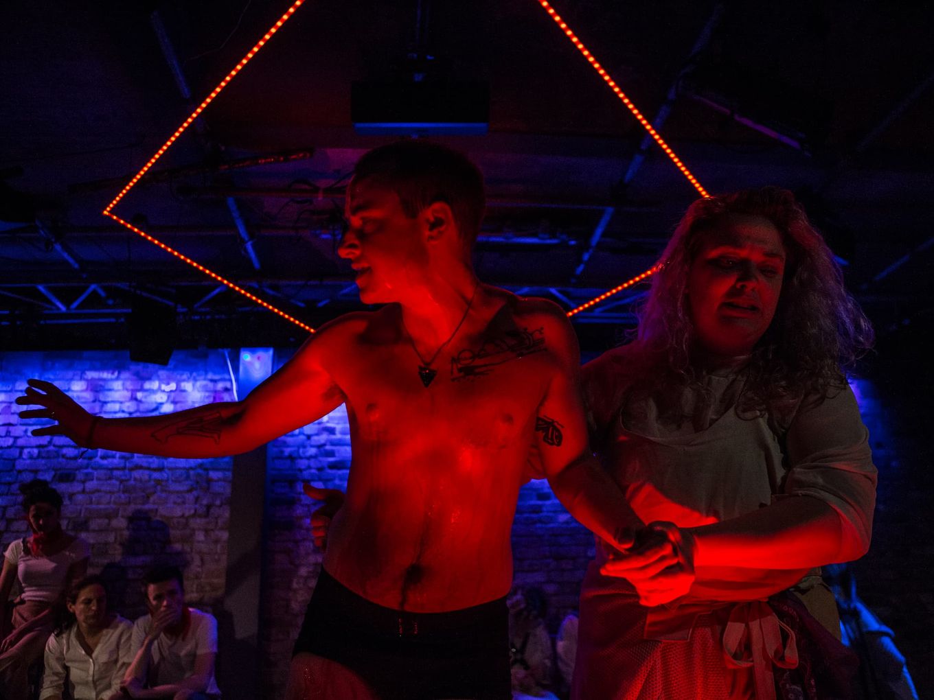 Under intense red light, a nearly naked man, dripping wet, is led away by a woman with long hair.